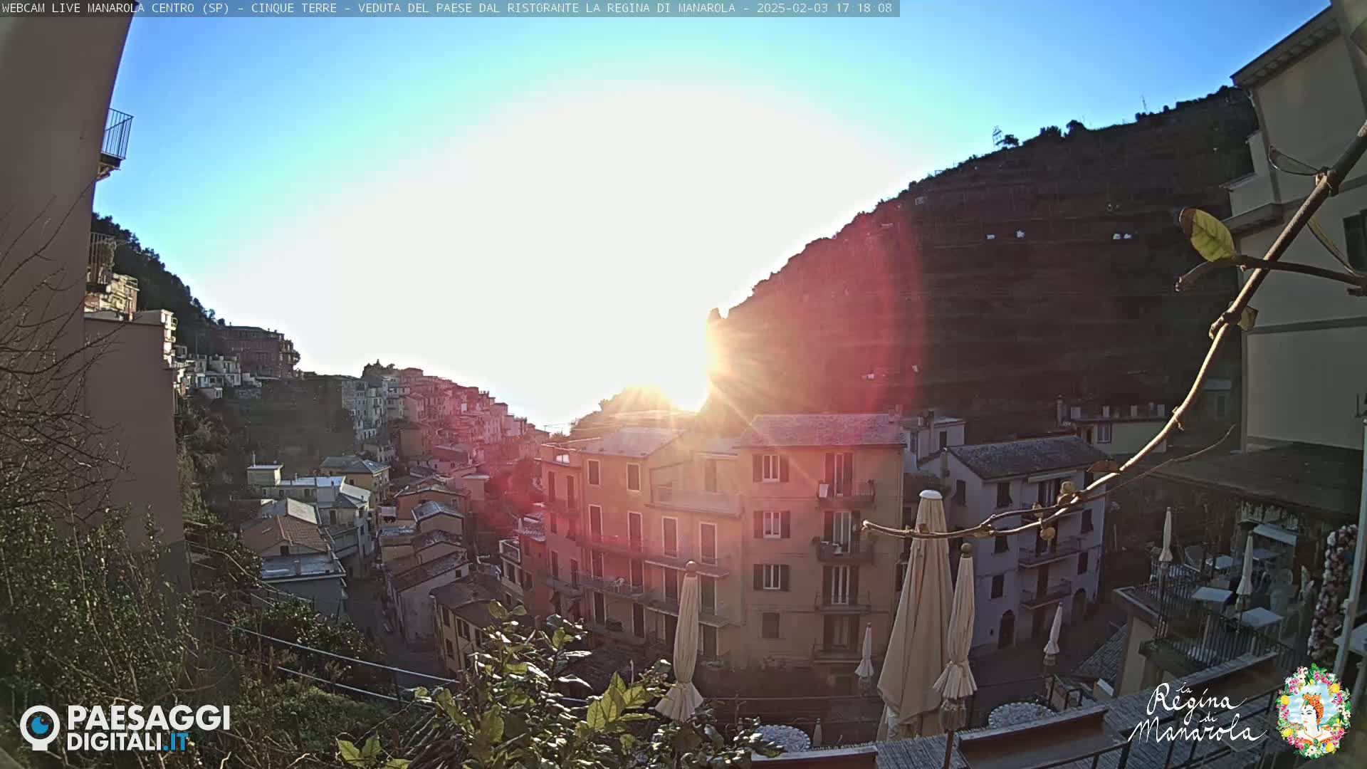 A sunlit view of a hillside town, with buildings cascading down a steep slope towards the sea, under a clear sky.