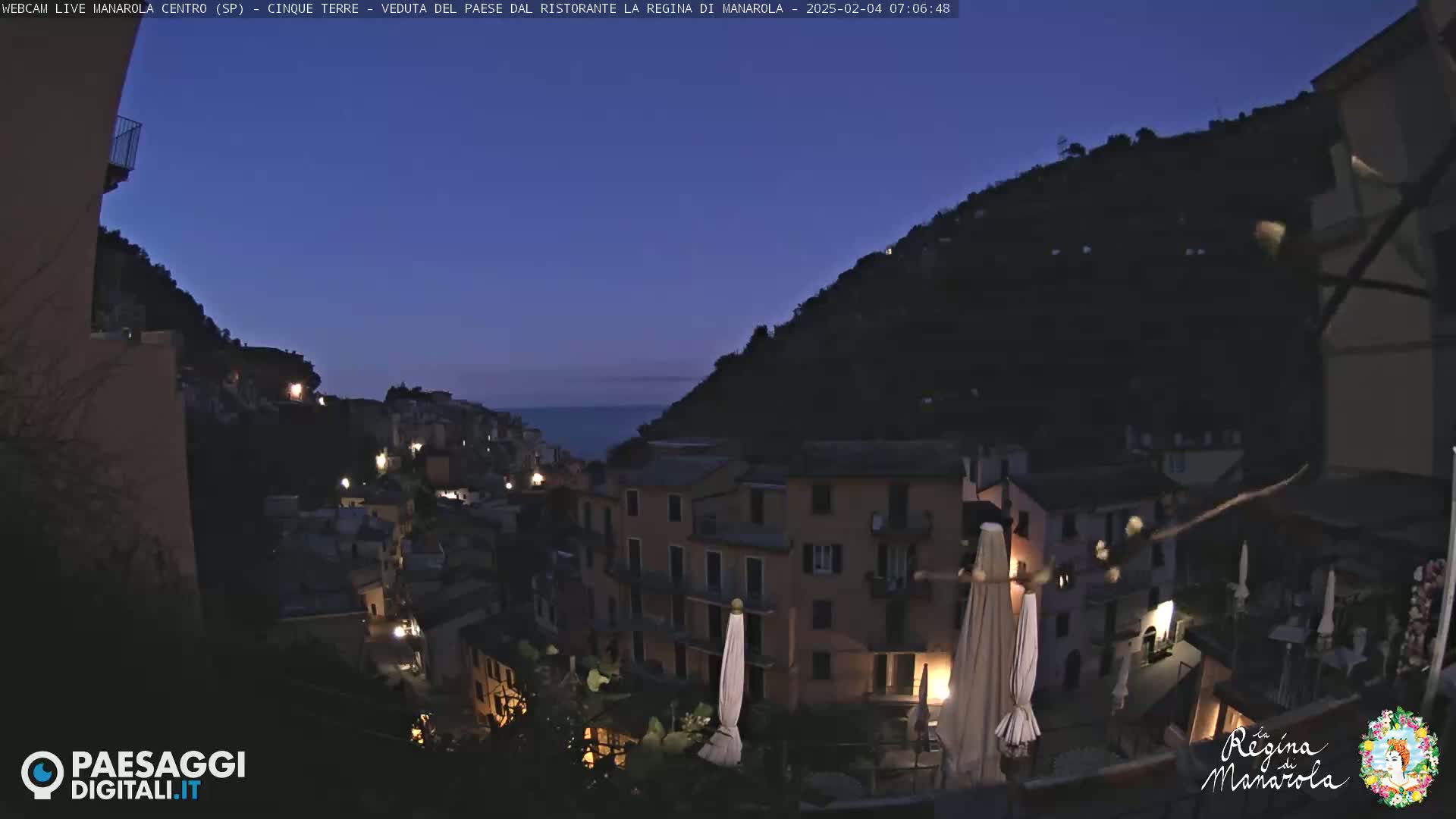 A twilight view of a hillside town overlooking a dark sea, with buildings illuminated by lights.