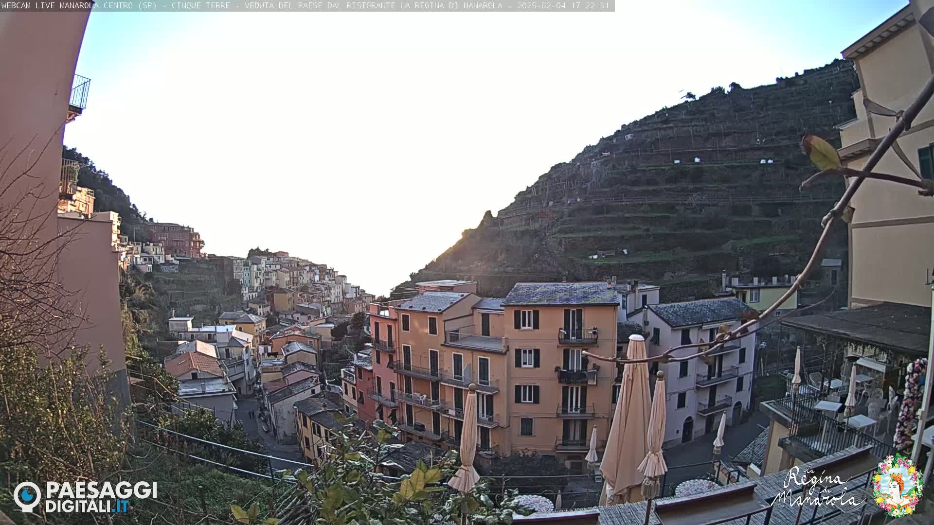 A sunlit coastal village is nestled into a hillside, terraced farms climbing the slopes behind multi-story buildings.
