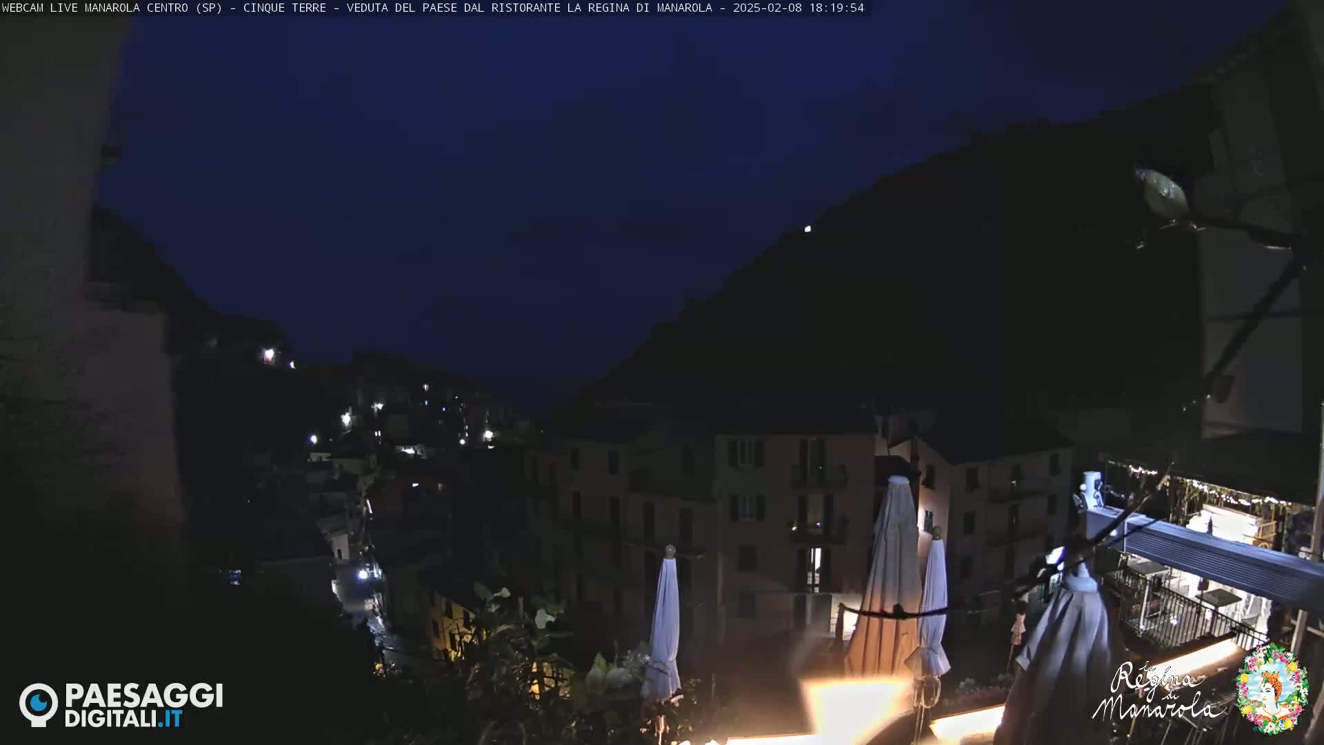 A nighttime view of a village nestled against a dark hillside, illuminated by scattered lights, with closed patio umbrellas in the foreground.