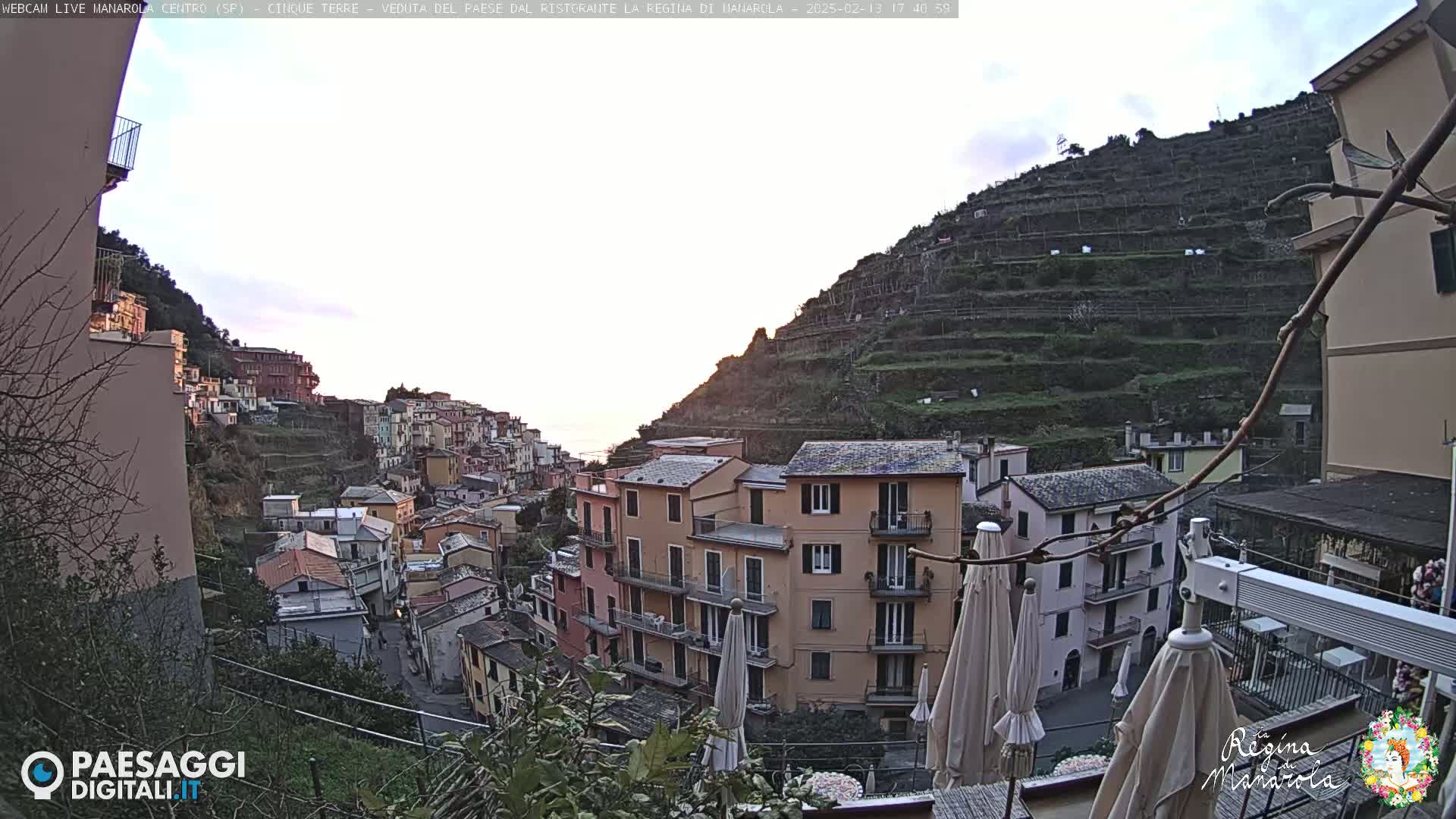 A hillside village with pastel-colored buildings, terraced vineyards, and patio umbrellas is seen under a mostly clear, twilight sky.