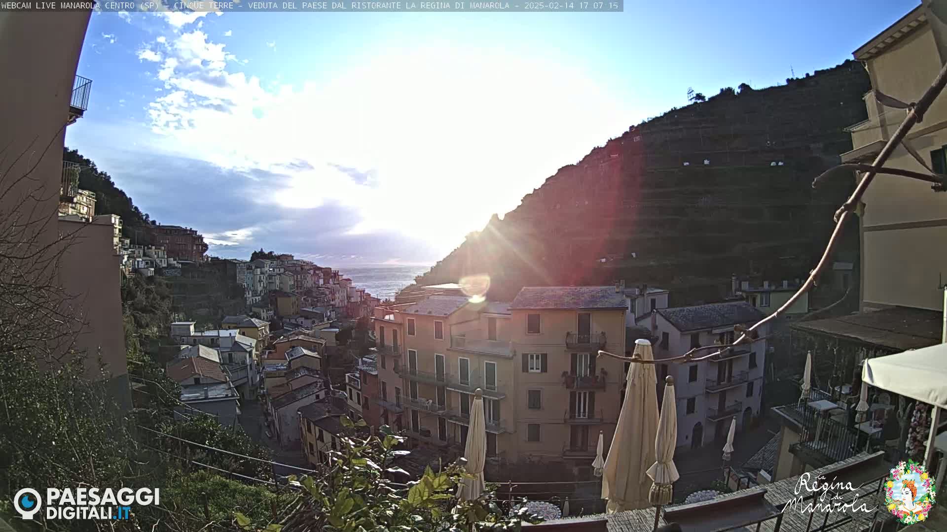 A sunlit coastal town nestled on a hillside, with buildings cascading down towards a calm sea under a partly cloudy sky.