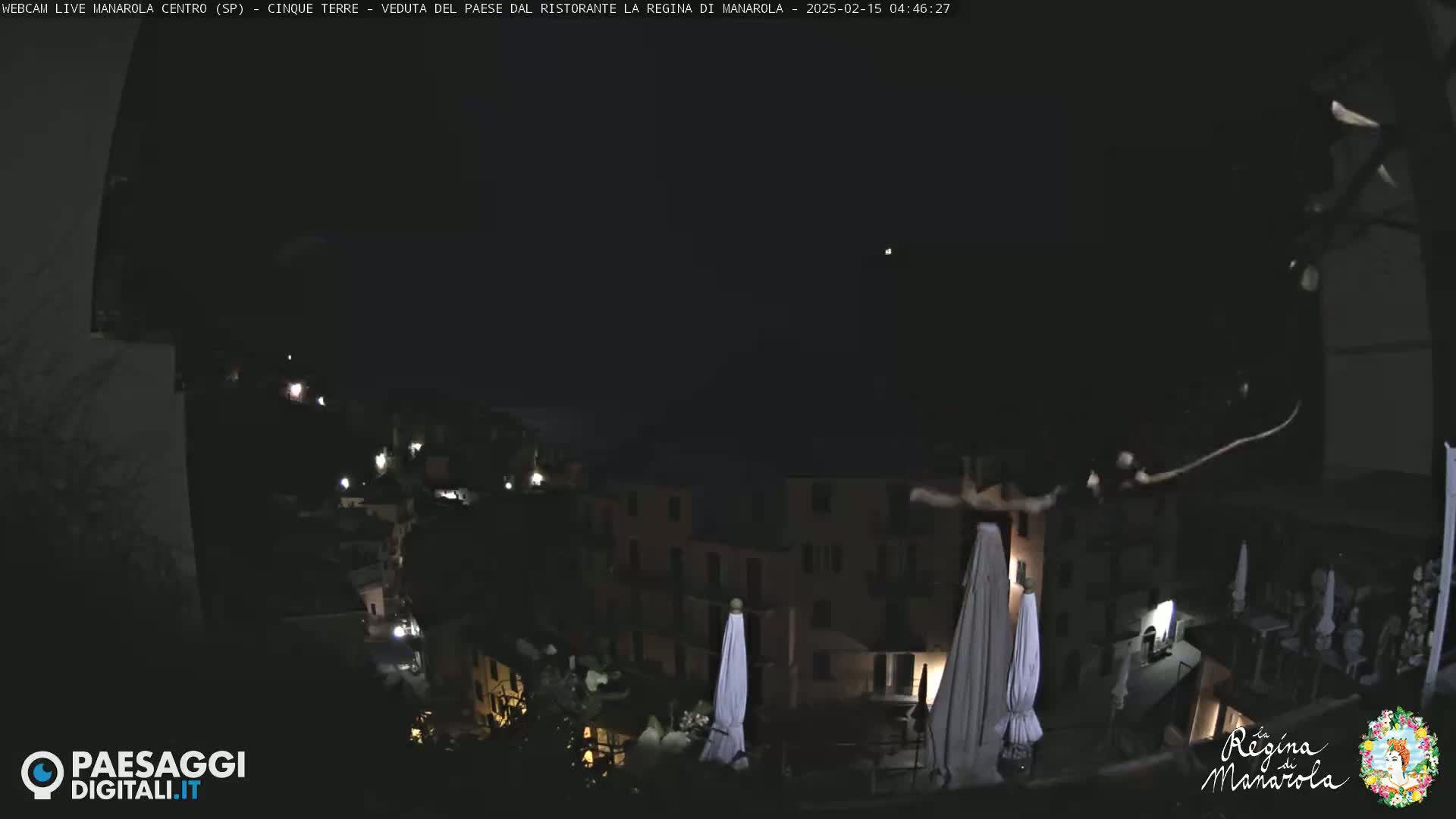 A nighttime view of a village illuminated by scattered lights, with closed umbrellas visible on a patio overlooking the scene.