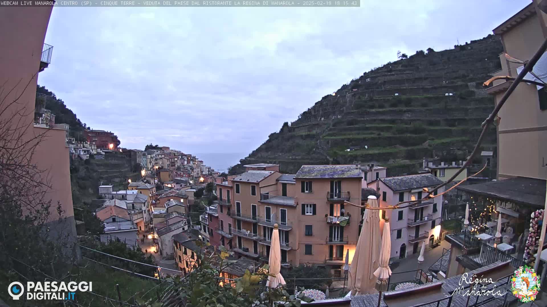 A twilight view of a hillside village with terraced fields, overlooking the sea under a cloudy sky.