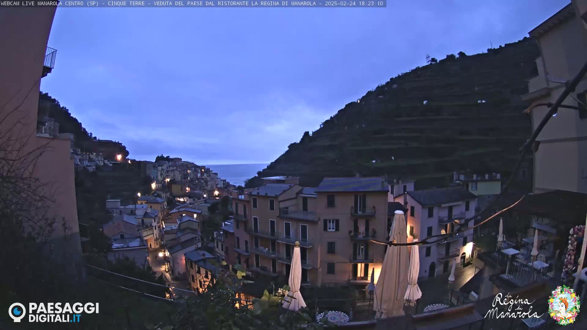 A twilight view of a hillside village with buildings lit up against a mostly clear, dark blue sky, overlooking a calm sea.