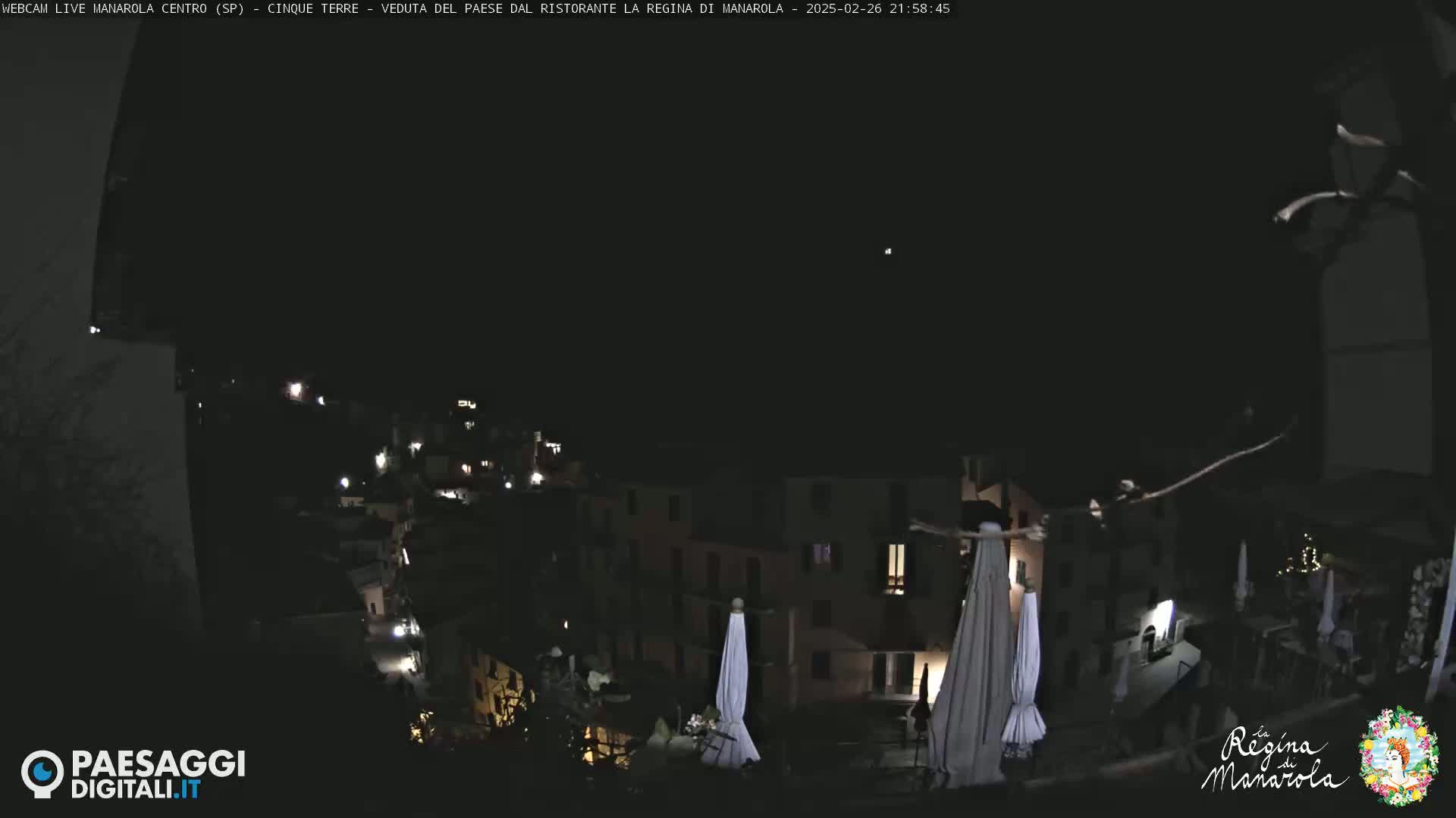 A nighttime, long shot of a village nestled on a hillside, with sparsely lit buildings and a few closed umbrellas visible.
