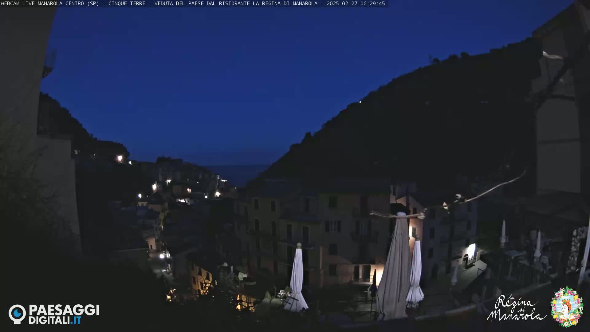 A nighttime view of a hillside village with buildings illuminated, overlooking a dark body of water under a clear, dark blue sky.