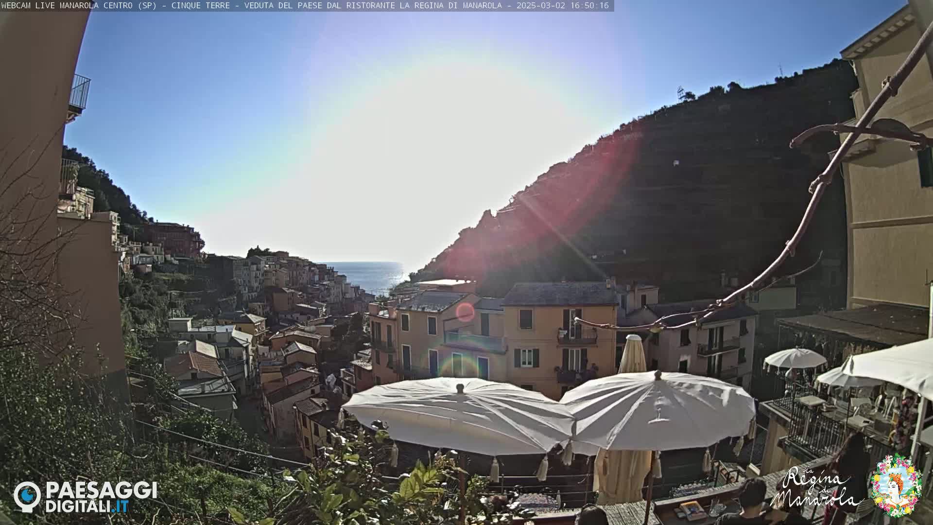 A sunny view of a colorful hillside village overlooking the sea, with white patio umbrellas in the foreground.