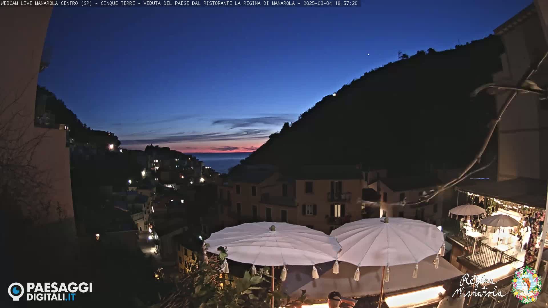 A twilight view of a coastal village nestled on a hillside, with the setting sun painting the sky with hues of pink and orange, and several white parasols on a patio in the foreground.