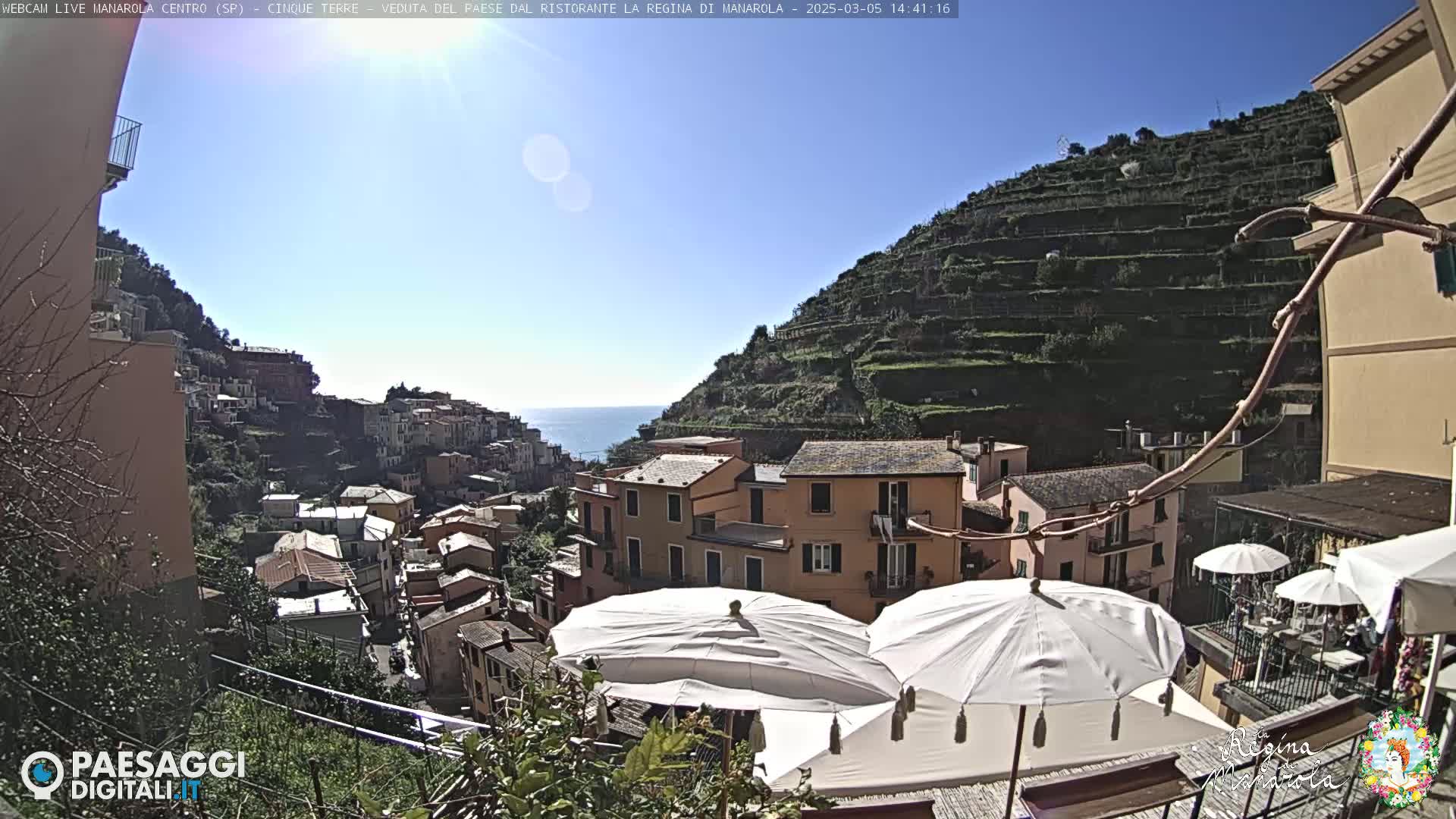A sunny, clear day overlooks a hillside village with terraced vineyards and buildings, and white patio umbrellas are in the foreground.