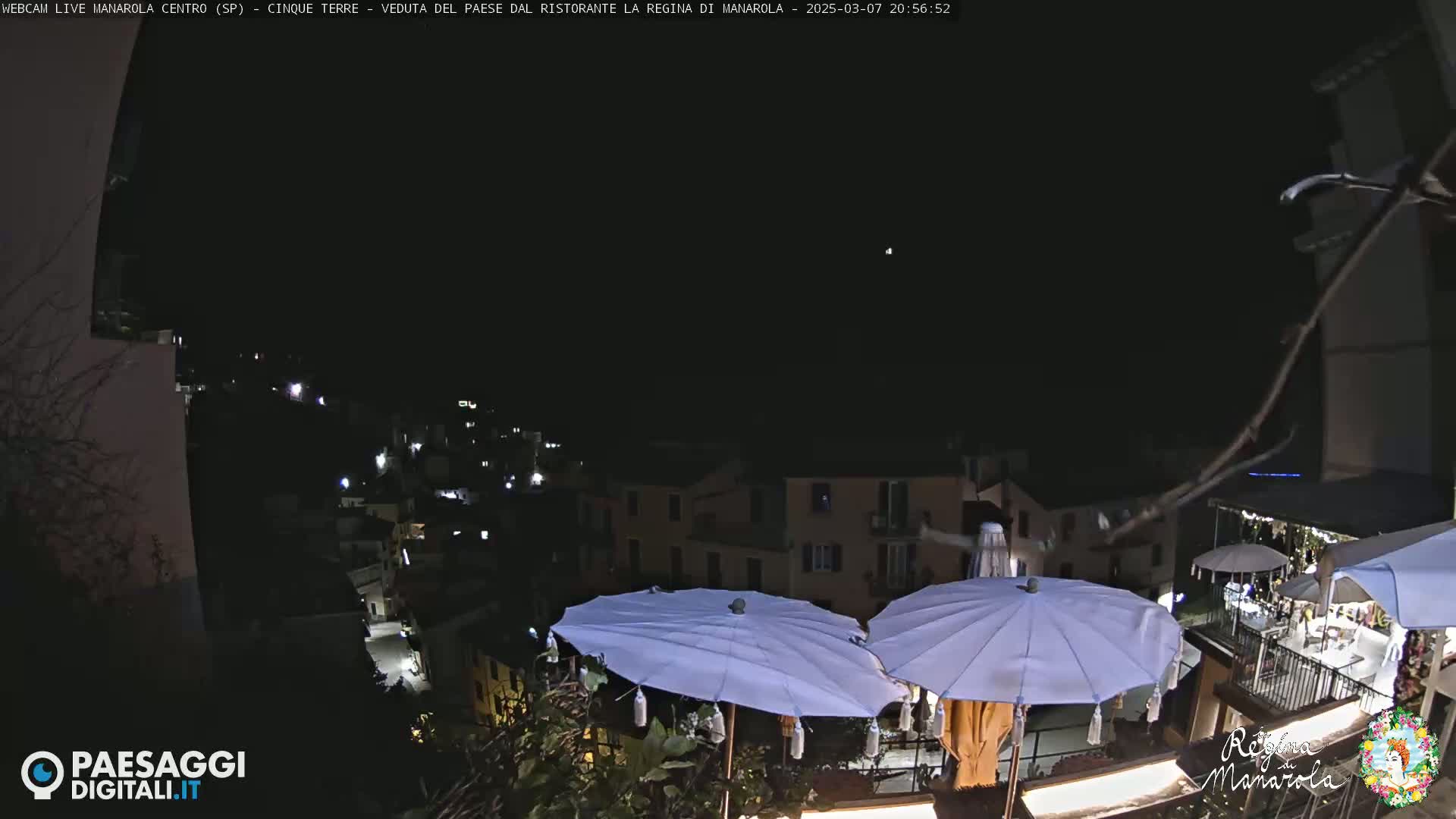 A nighttime view of a village nestled on a hillside, illuminated by scattered lights, with several closed white umbrellas on a patio in the foreground under a clear, dark sky.