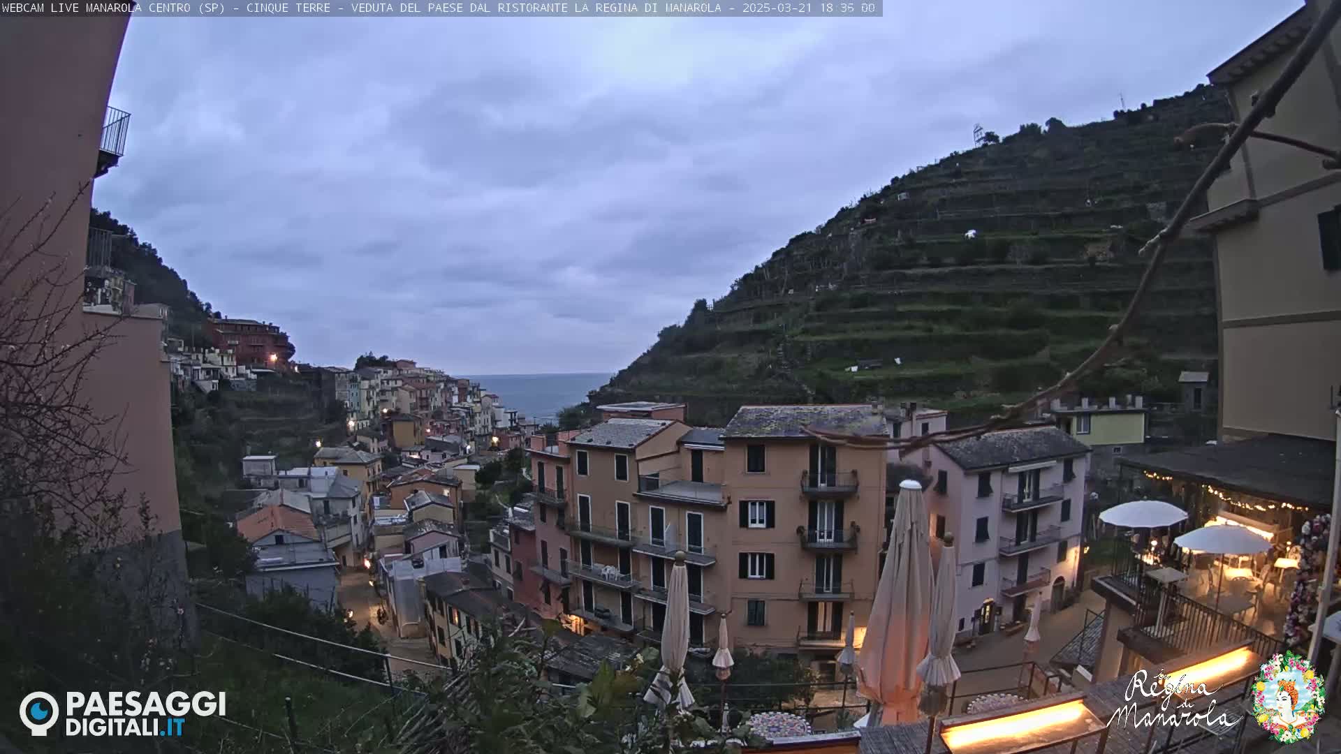 A twilight view of a coastal village nestled on a hillside, with terraced gardens climbing the slopes above, under a cloudy sky.