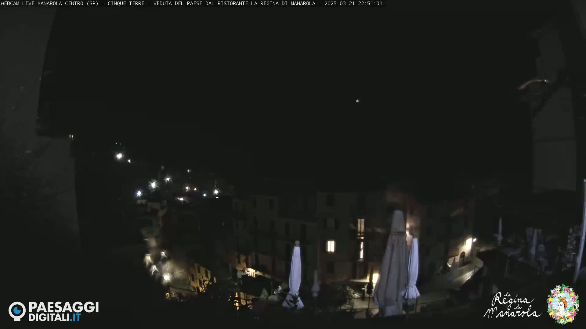 A nighttime view of a village, showing buildings with some lights on, and closed umbrellas on a patio.
