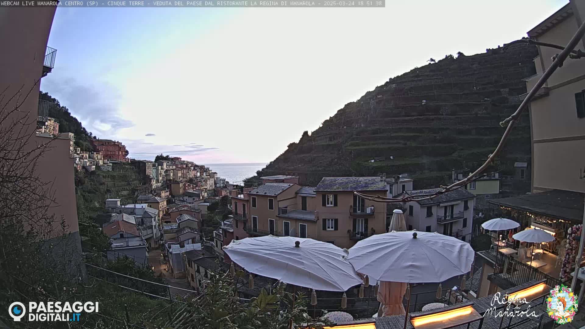 A coastal village clings to a hillside overlooking a calm sea under a mostly clear, twilight sky; two white umbrellas sit on a patio overlooking the scene.