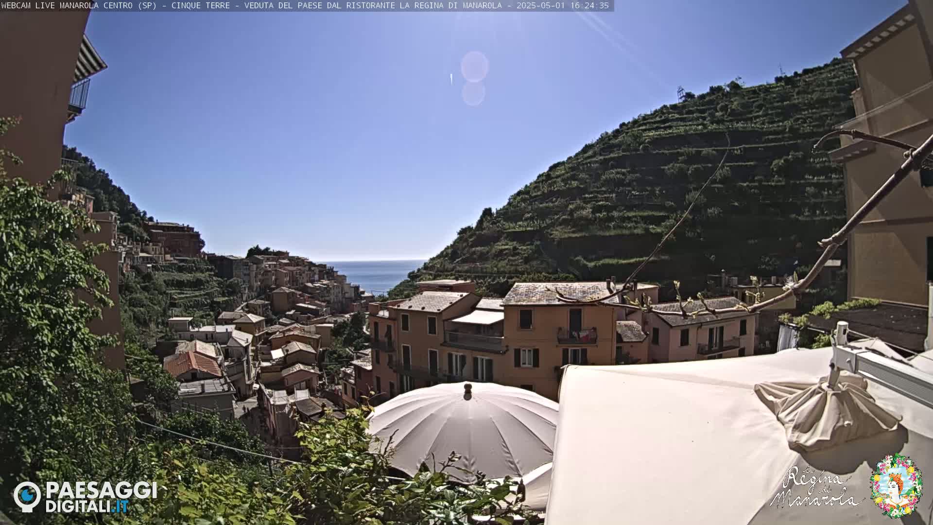 A sunny, seaside village clings to a terraced hillside, with an umbrella partially obscuring the view from a patio.