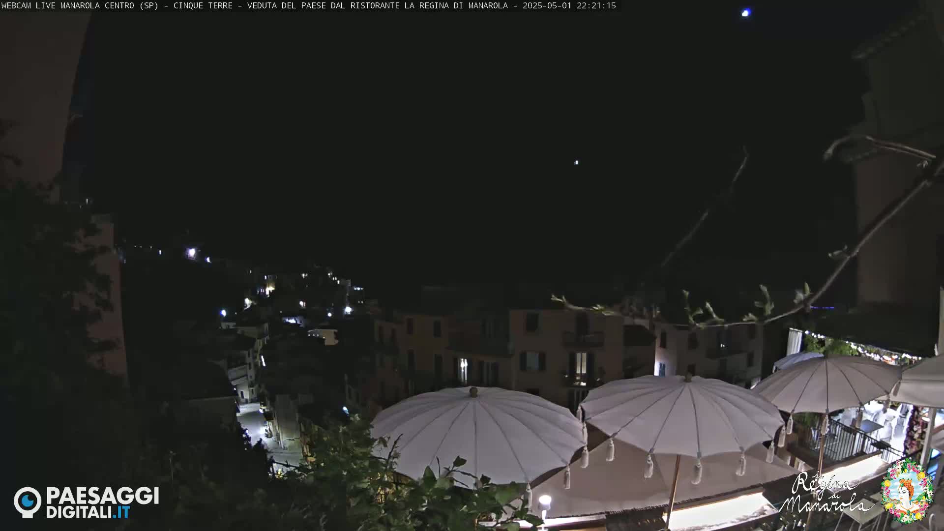 A nighttime view of a village nestled on a hillside, illuminated by scattered lights, with several white parasols in the foreground under a clear, dark sky.