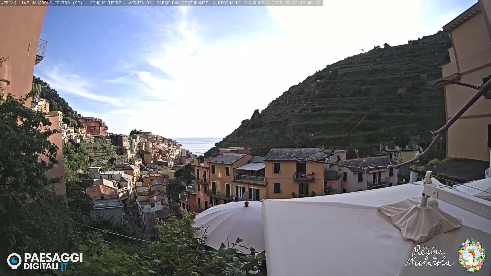 A coastal village clings to a steep hillside under a mostly clear sky, partially obscured by an umbrella and other patio furniture.