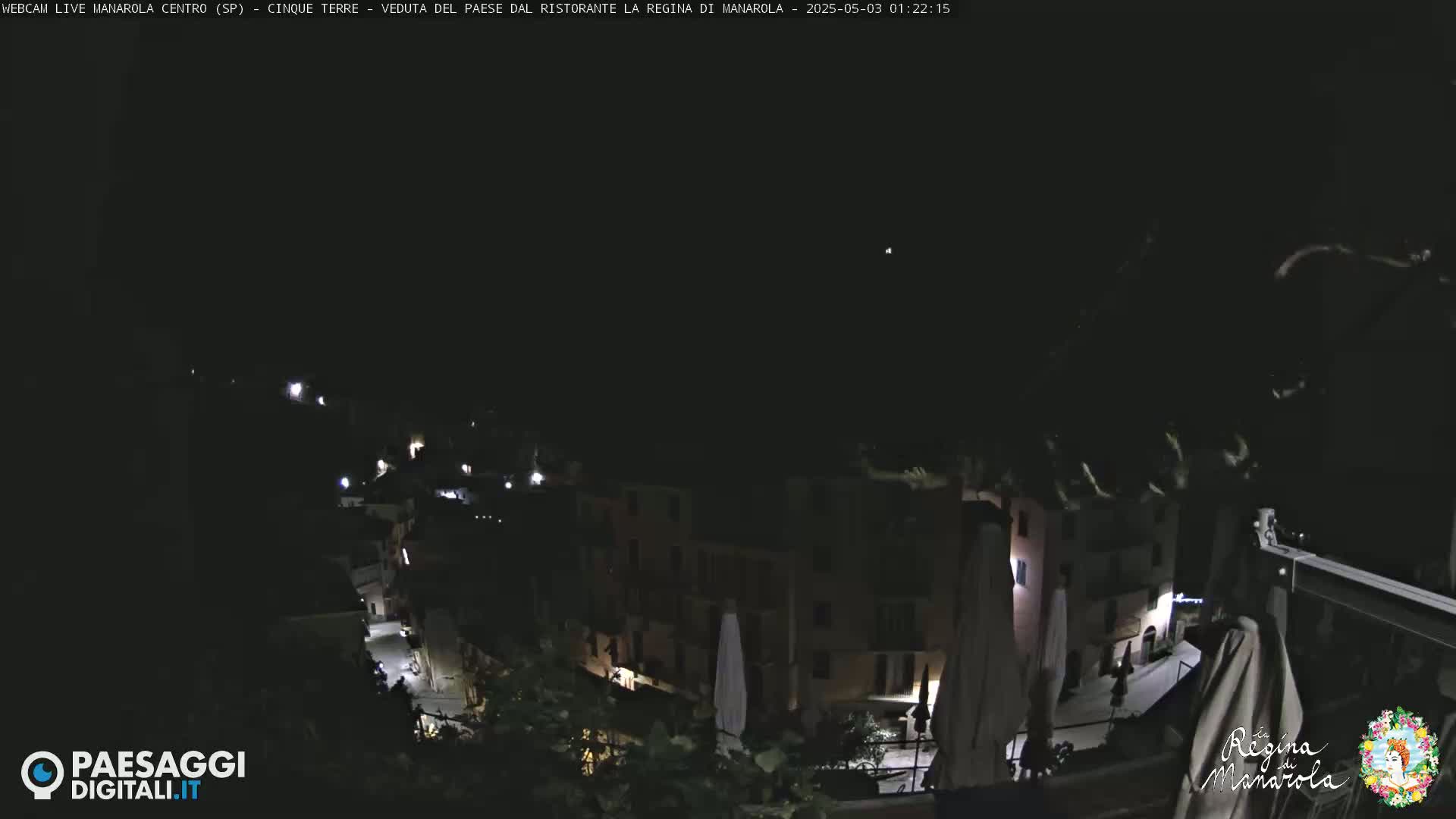 A nighttime view of a village nestled on a hillside, with some lights visible from buildings and a few faint lights on the street below.