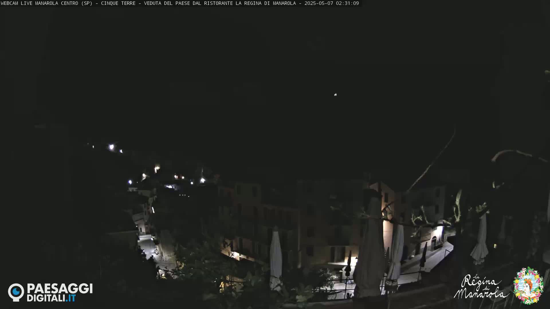 A nighttime, high-angle view of a dimly lit town nestled on a hillside, with a few lights visible along the streets and buildings.