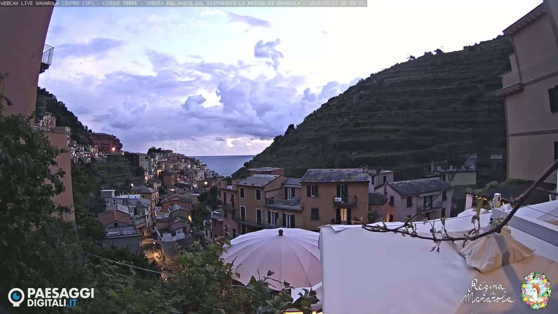 A colorful hillside village overlooks a calm sea under a partly cloudy, dusky sky.