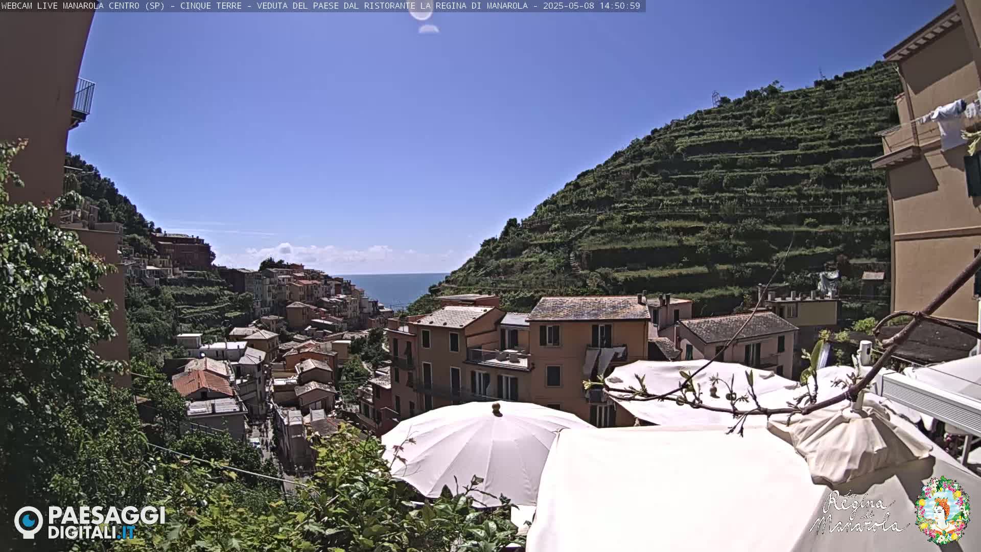 A sunny view of a hillside town overlooking the ocean, with terraced gardens and white patio umbrellas in the foreground.