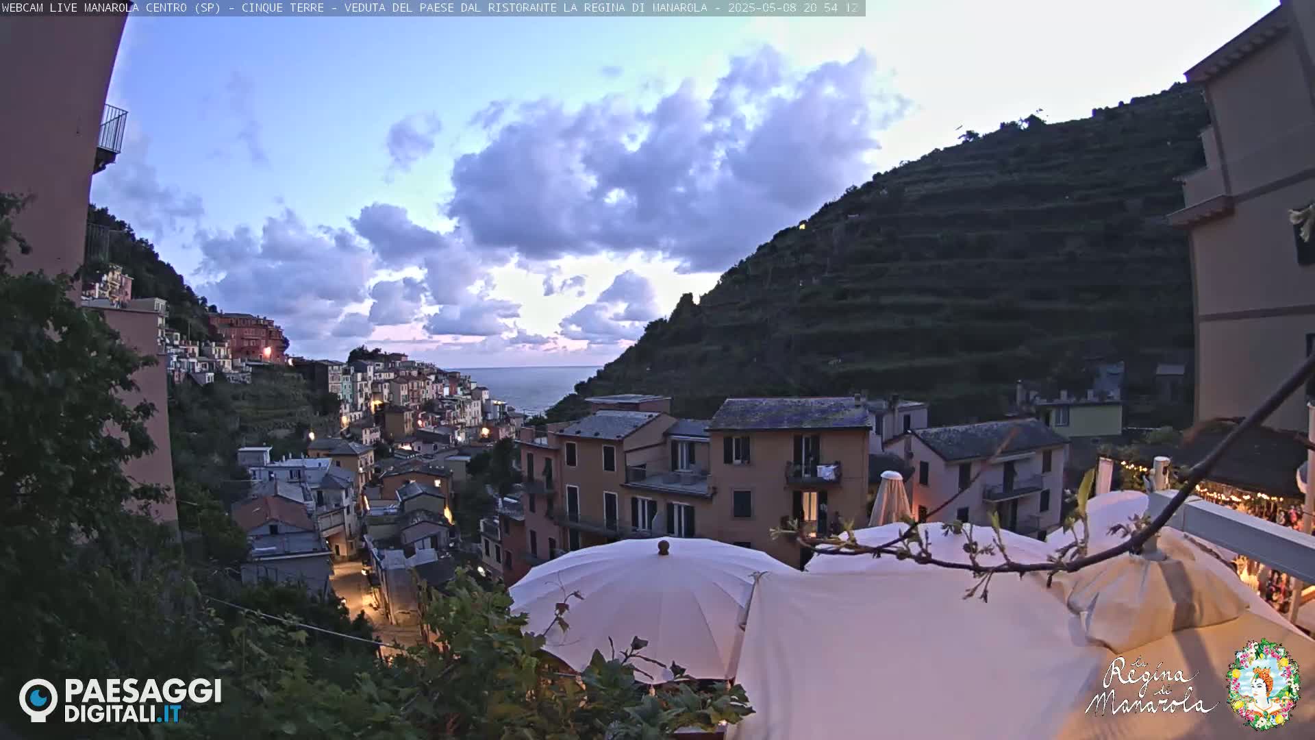 A twilight view of a colorful hillside village overlooking a calm sea under a partly cloudy sky.