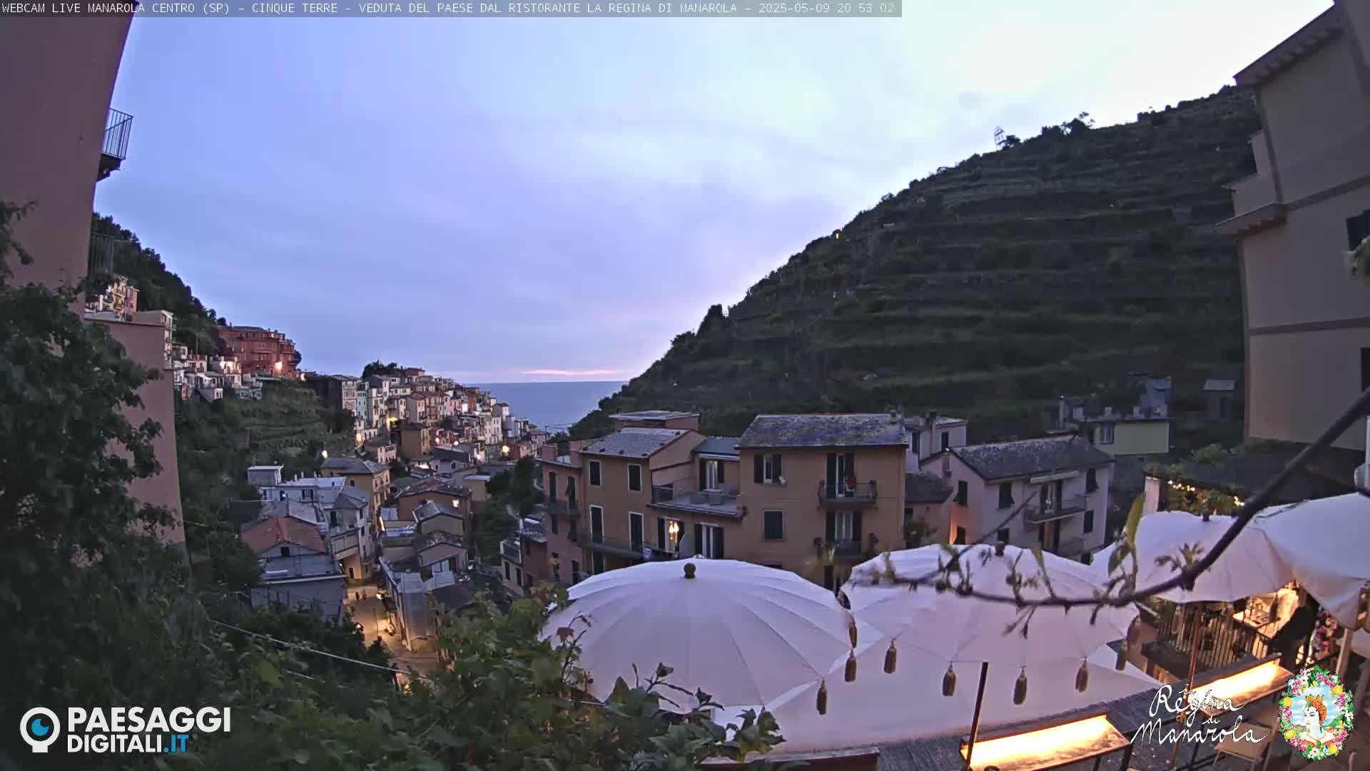 A twilight view of a hillside village with buildings lit up, terraced gardens, and several outdoor parasols.