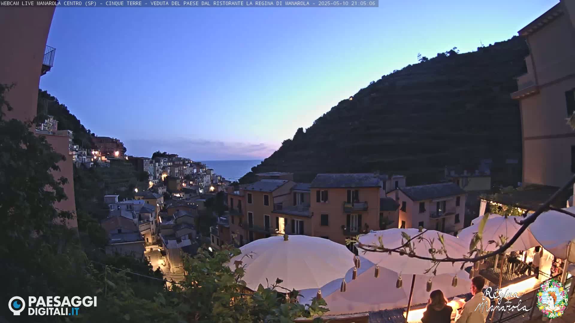 A twilight view of an Italian hillside village with illuminated buildings, overlooking a calm sea, features several outdoor umbrellas at a restaurant.
