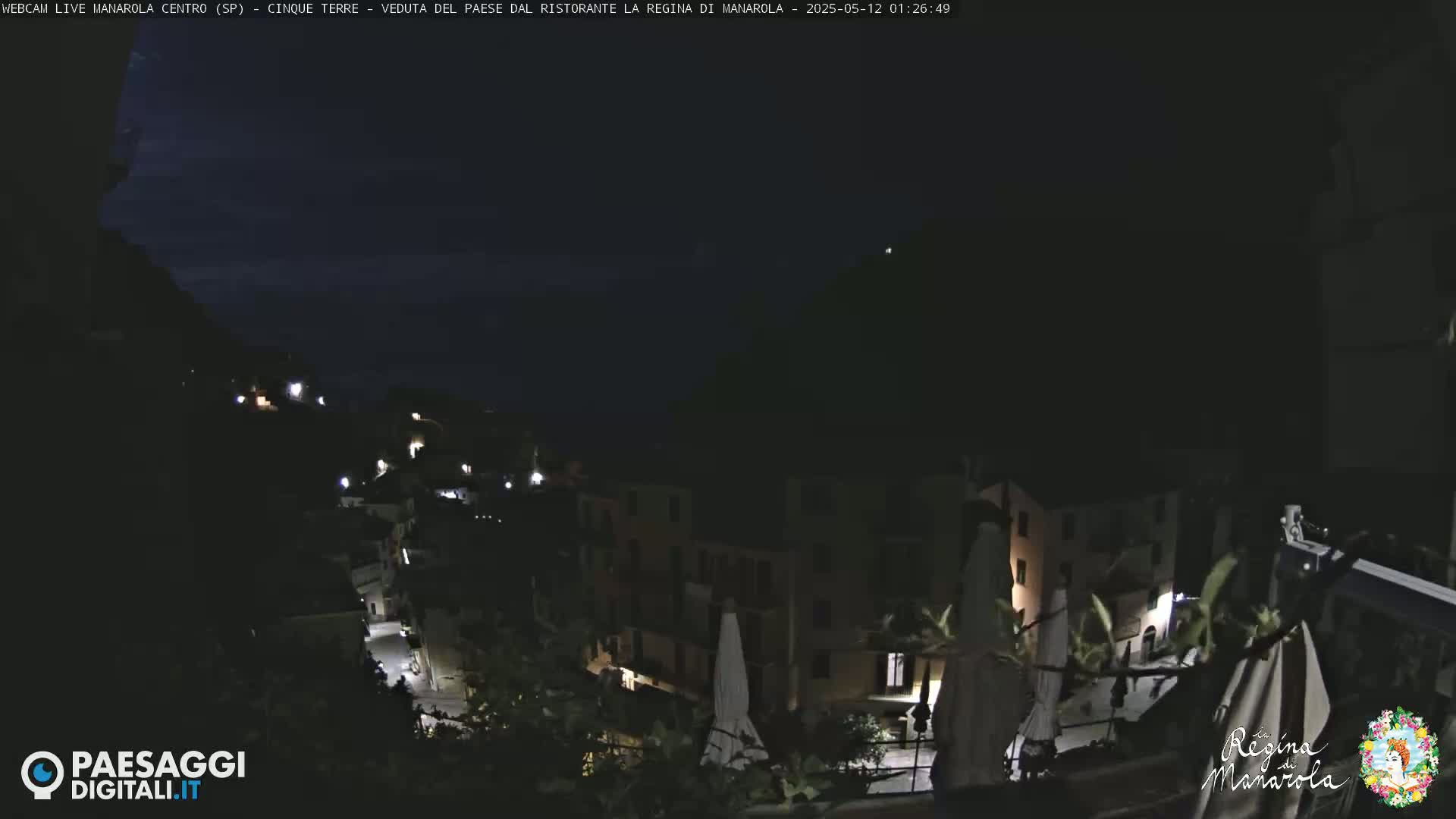 A nighttime view of a dimly lit hillside town with a few lights visible, under a dark, mostly clear sky.