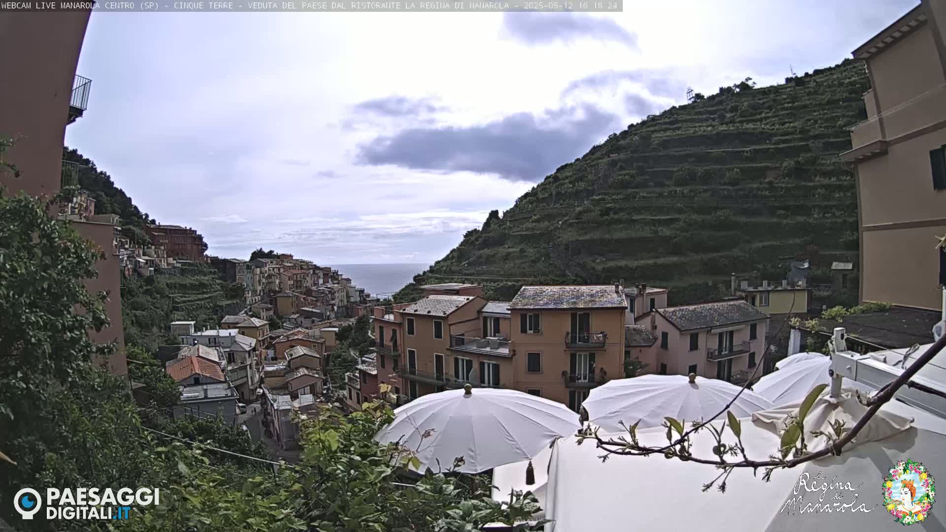 Overcast skies loom over a picturesque coastal village nestled on a steep hillside, terraced gardens cascading down to the sea, with patio umbrellas partially obscuring the view.