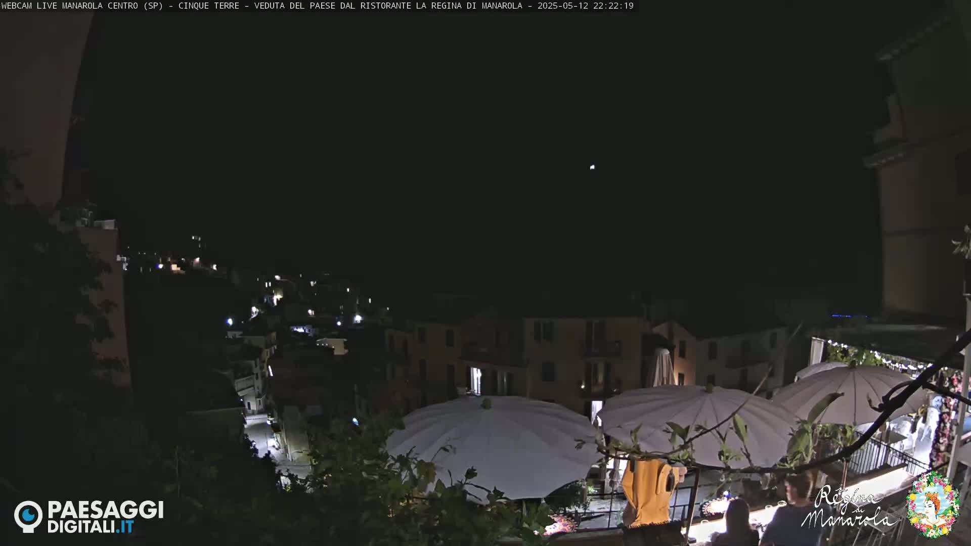 A nighttime view of a village nestled on a hillside, with outdoor patio umbrellas illuminated under a clear, dark sky.