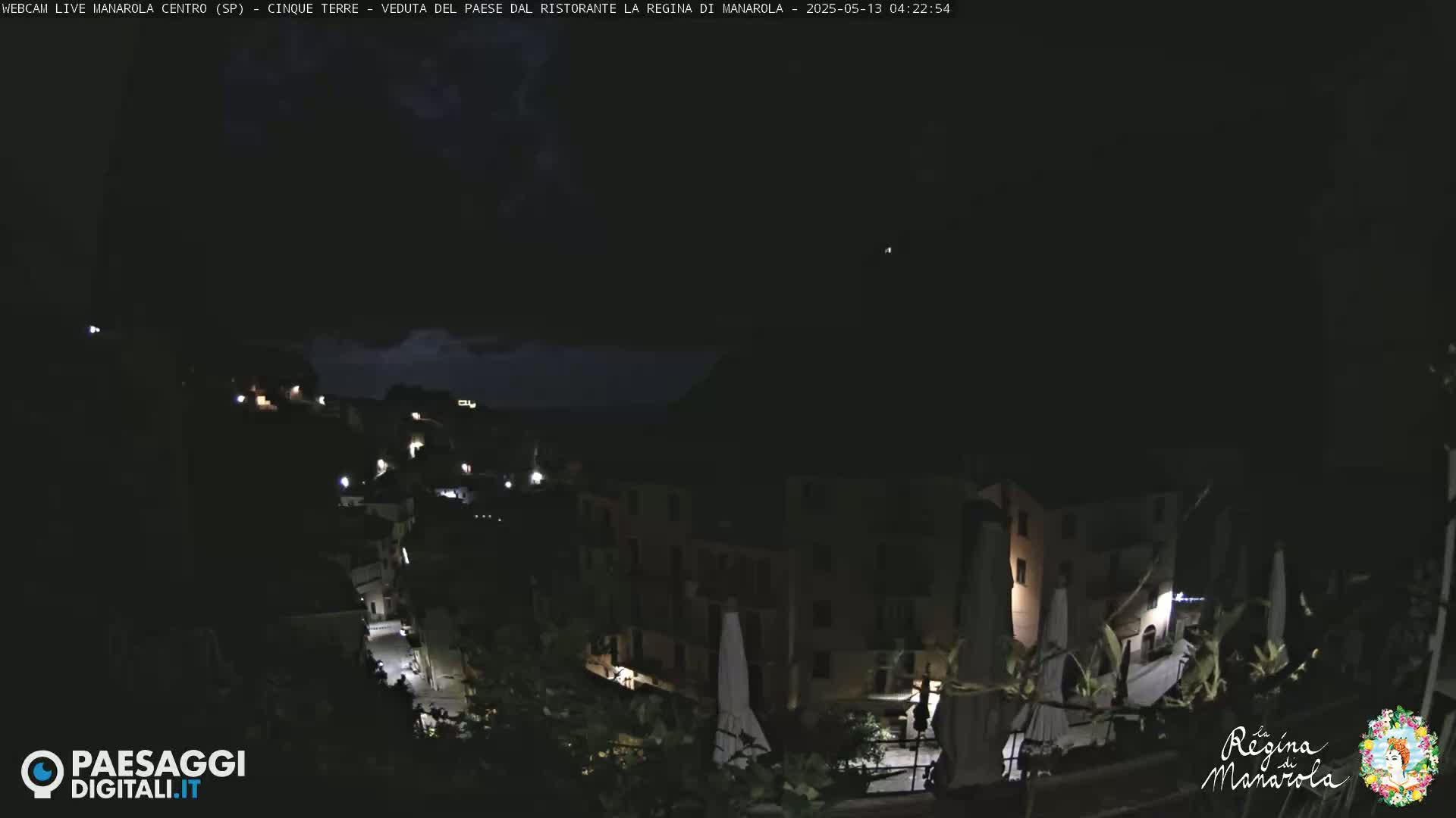 A nighttime view of a dimly lit hillside town with a few lights visible, under a dark, possibly cloudy sky.
