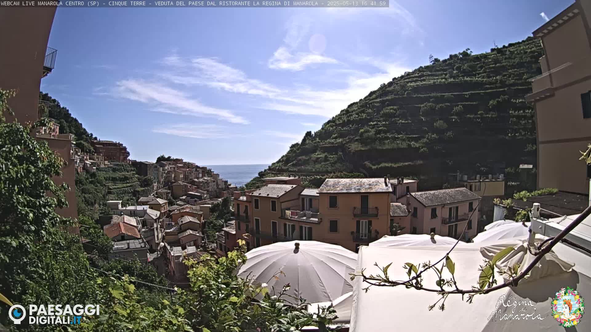 A sunny day overlooking a colorful hillside town nestled against a terraced mountainside, with the sea visible in the distance, and several patio umbrellas in the foreground.