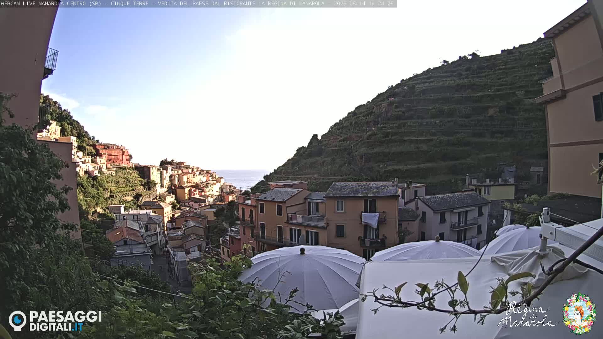 A coastal village nestled on a hillside overlooking the sea under a clear sky, with several white patio umbrellas in the foreground.