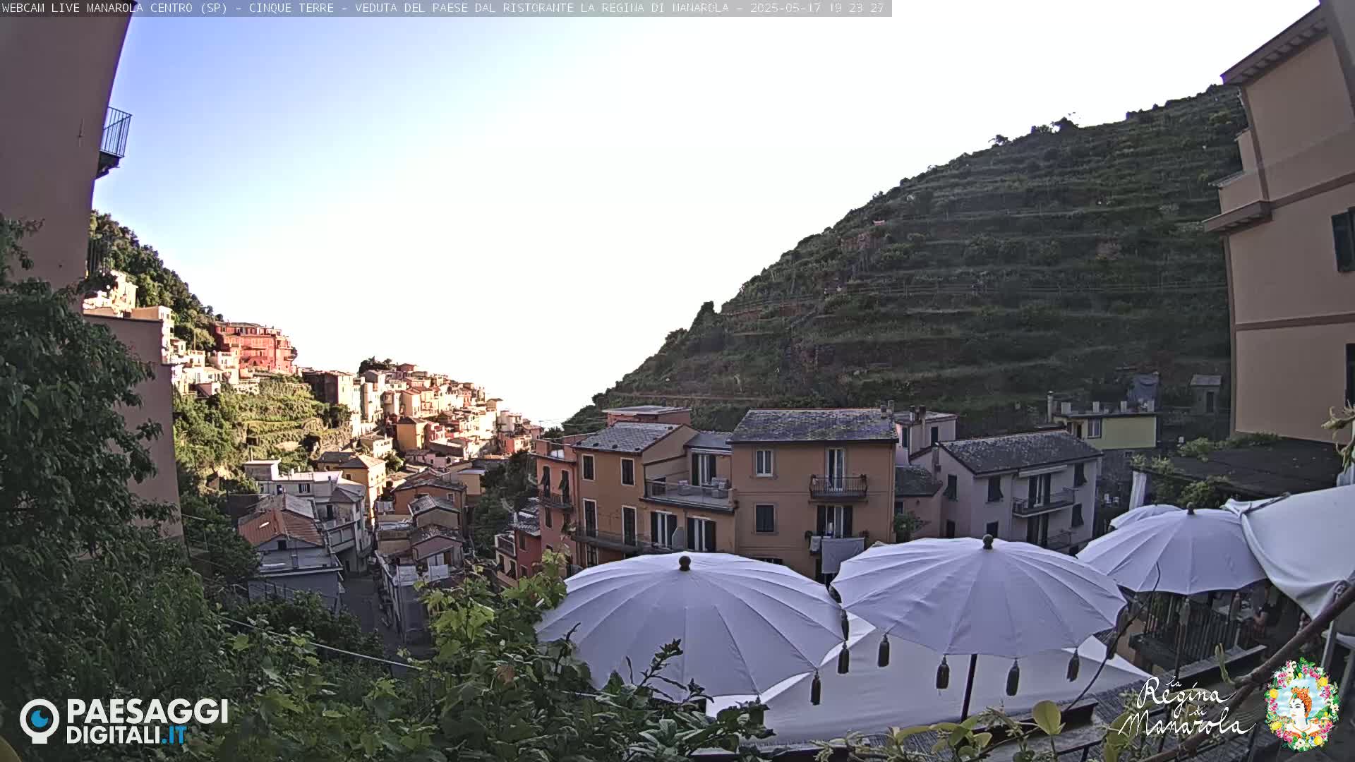 A hillside village with colorful buildings is seen from a patio with white umbrellas, under a clear sky.