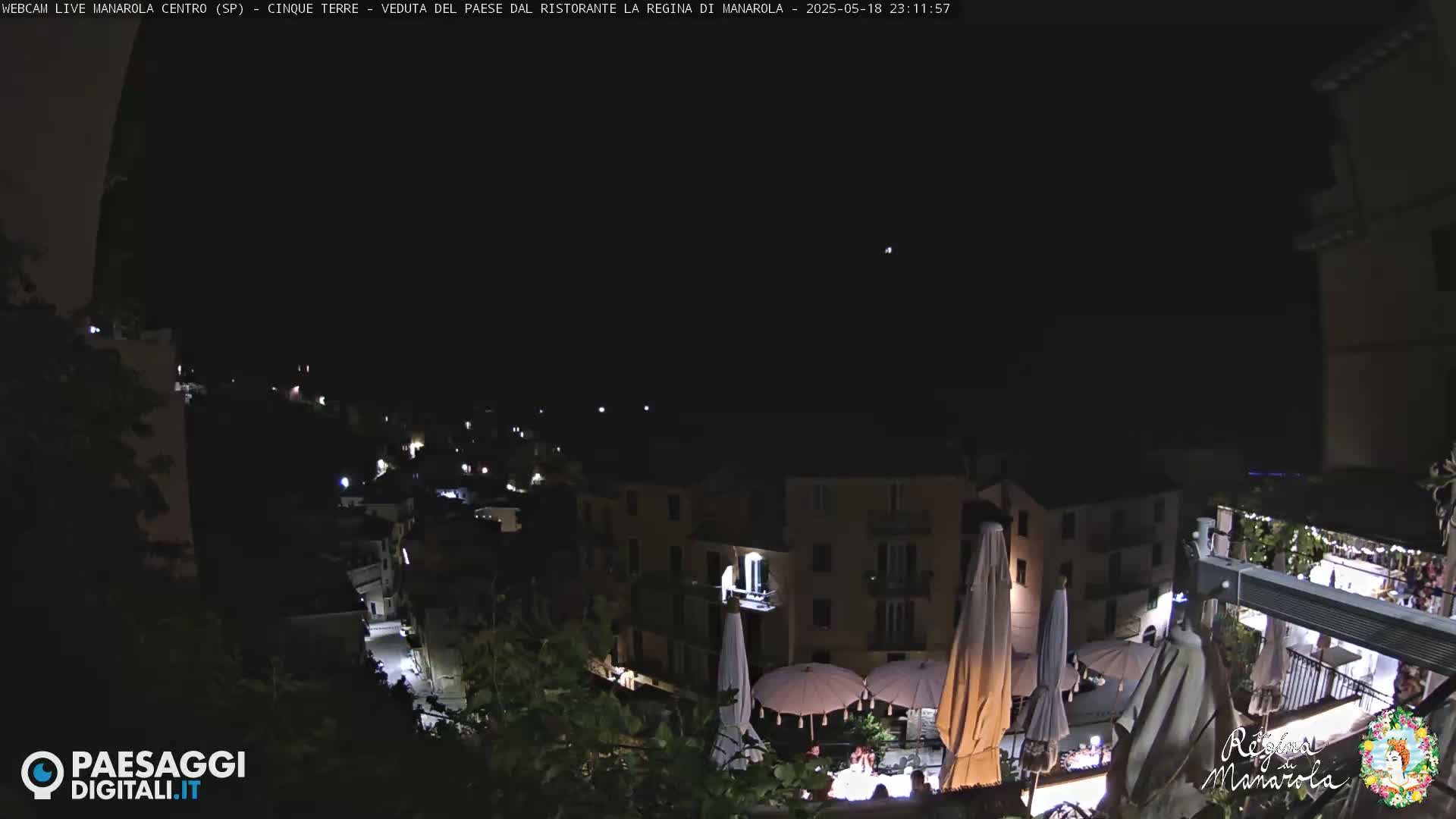 A nighttime, clear-sky view of a village, showing buildings with illuminated windows and outdoor patio umbrellas.