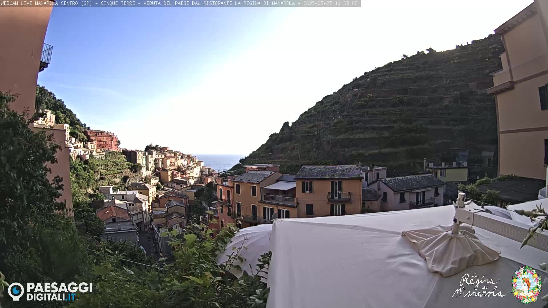 A clear, sunny day overlooks a picturesque Italian coastal village nestled on a hillside, with the sea visible in the distance.