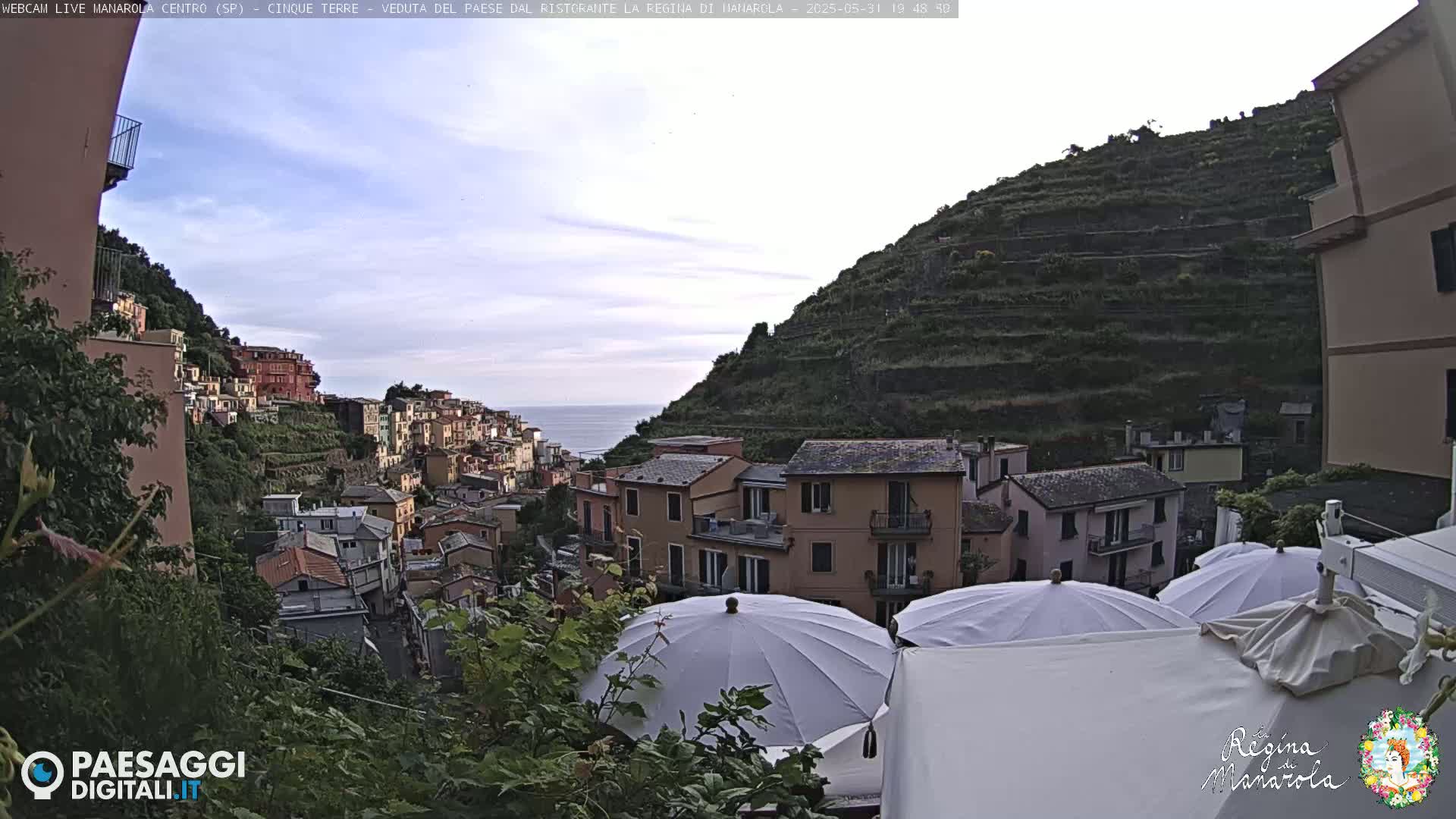 Overlooking a coastal village built into a hillside, under a mostly clear sky, are several closed white umbrellas.