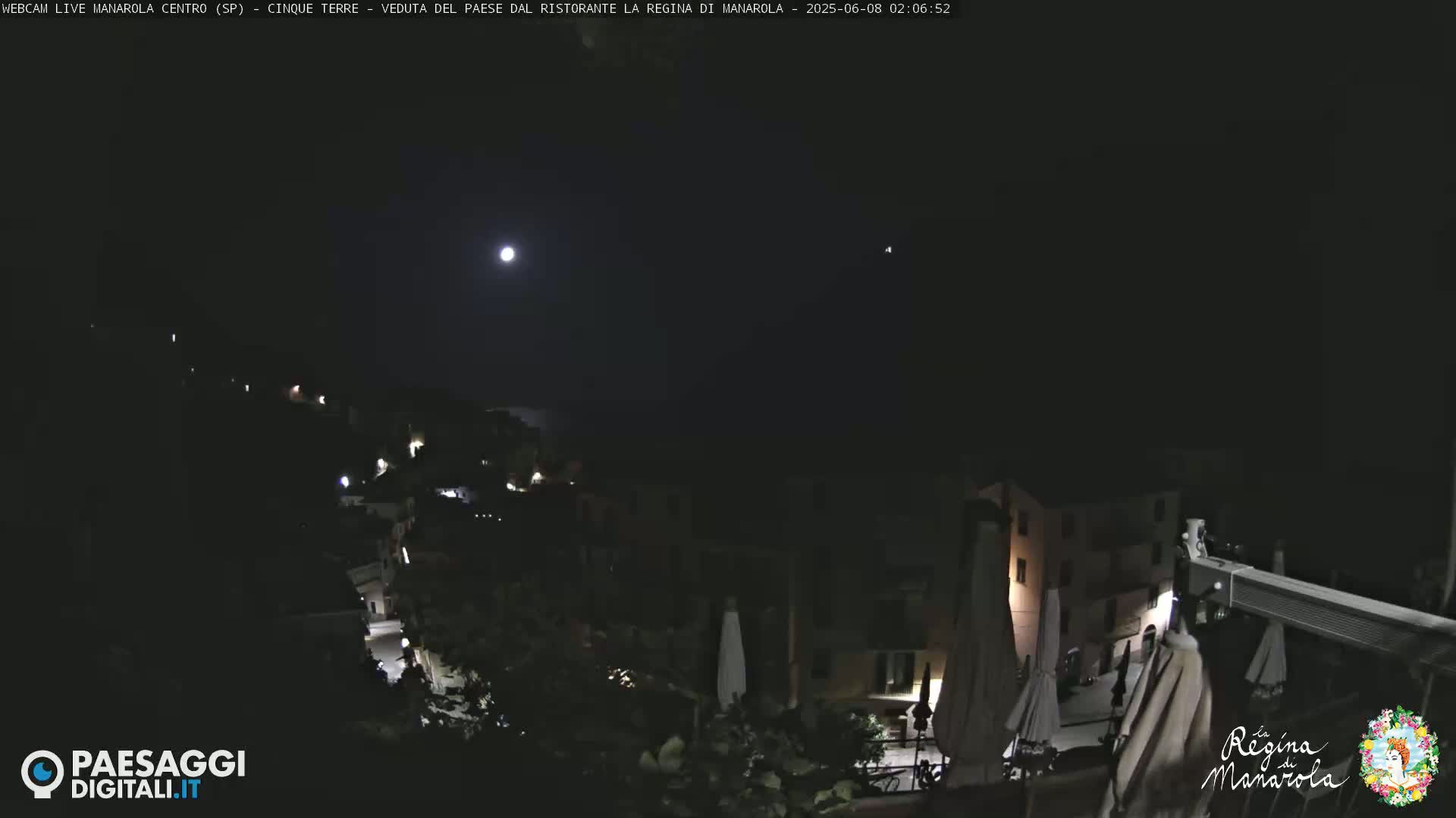 A nighttime view of a village, illuminated by lights and a bright moon, under a clear sky.