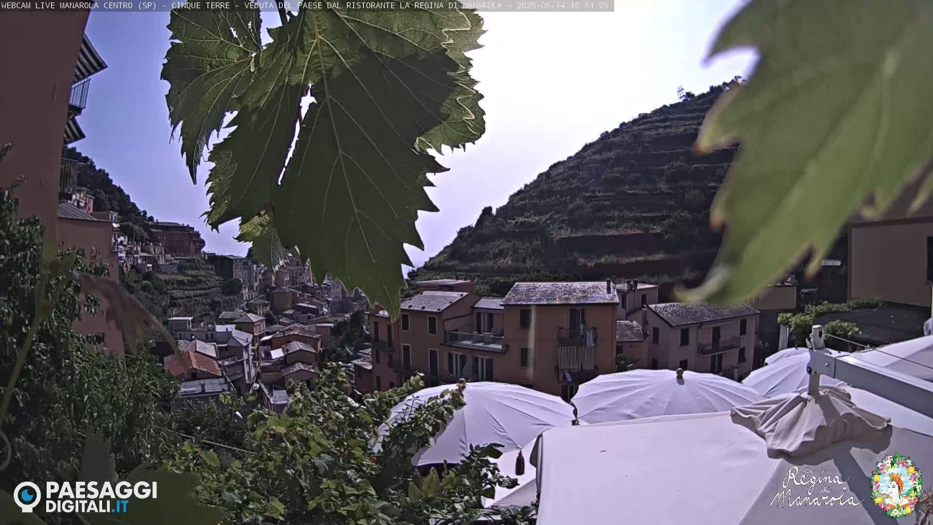 A hillside village with terracotta-roofed buildings is partially obscured by green foliage and white patio umbrellas under a mostly clear sky.