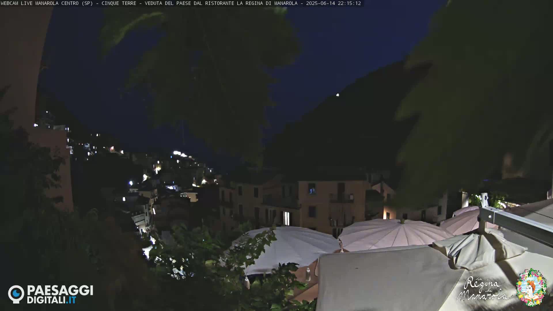 A hillside village with terracotta-roofed buildings is partially obscured by green foliage and white patio umbrellas under a mostly clear sky.