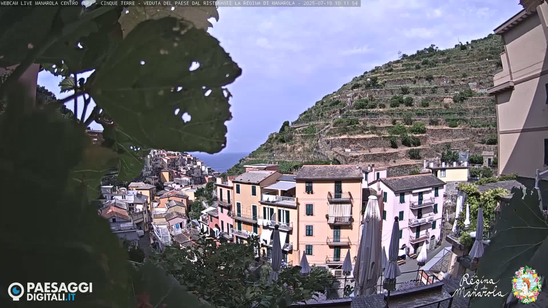 A coastal village with pastel-colored buildings climbs a terraced hillside under a mostly sunny sky, partially obscured by leafy vegetation in the foreground.