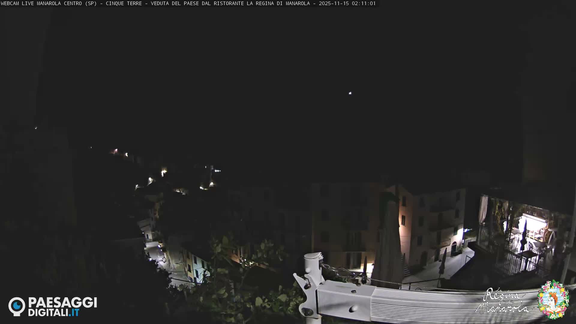 A clear, dark night sky with a single bright light overlooks a hillside town, where buildings and streets are softly illuminated by numerous artificial lights.