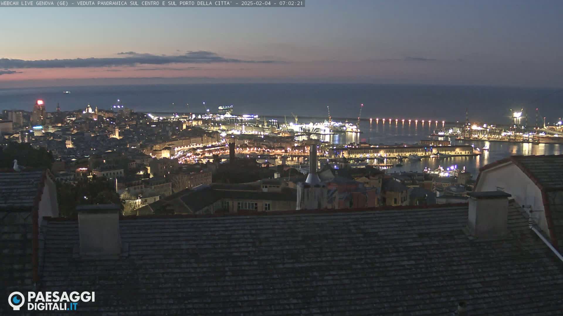 A nighttime panoramic view of a city and harbor, illuminated by lights, with a calm sea visible in the background under a twilight sky.