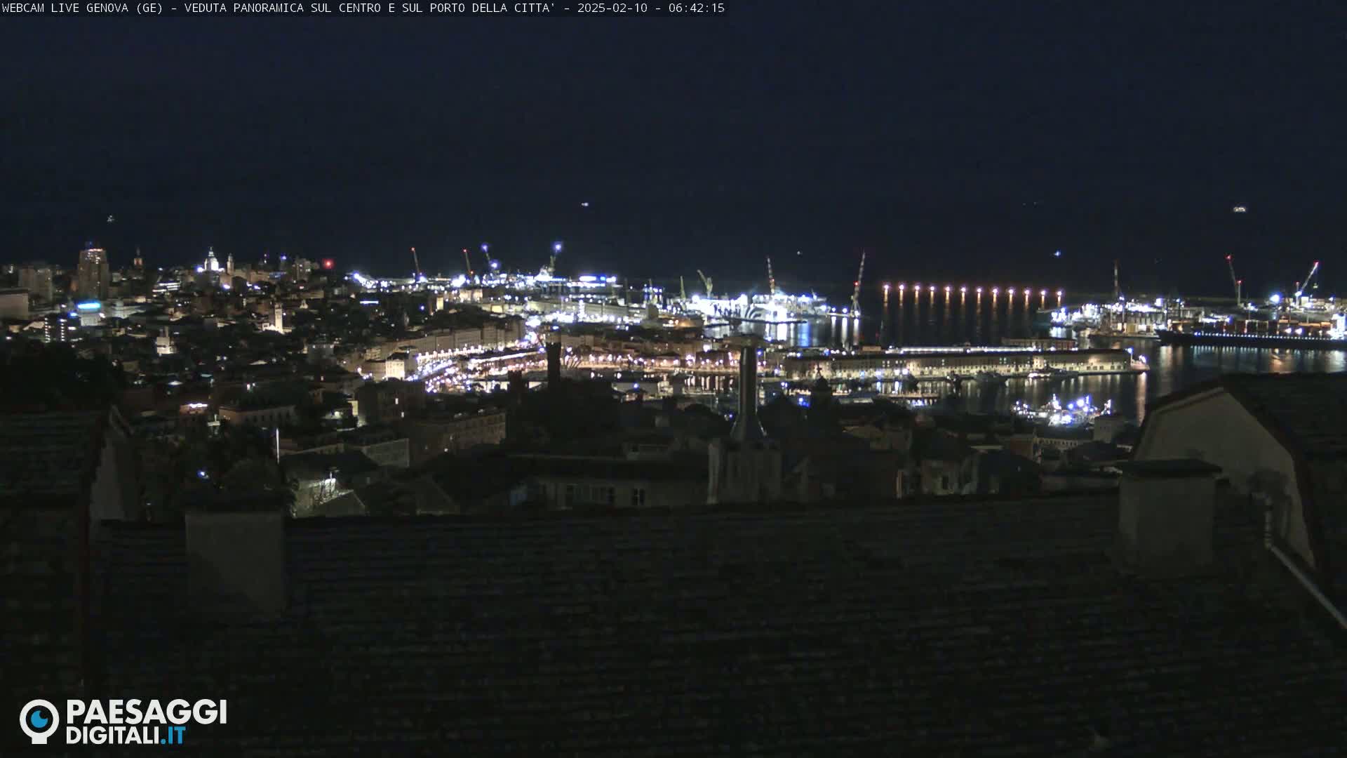 A nighttime, panoramic view of a city's illuminated harbor and buildings, extending to a dark coastline.