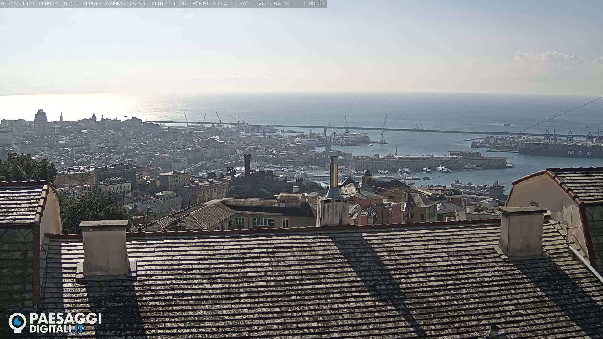 A panoramic view of a city and harbor, under a clear, sunny sky, shows a sprawling cityscape extending to a busy port filled with ships and cranes, with the calm sea visible in the distance.