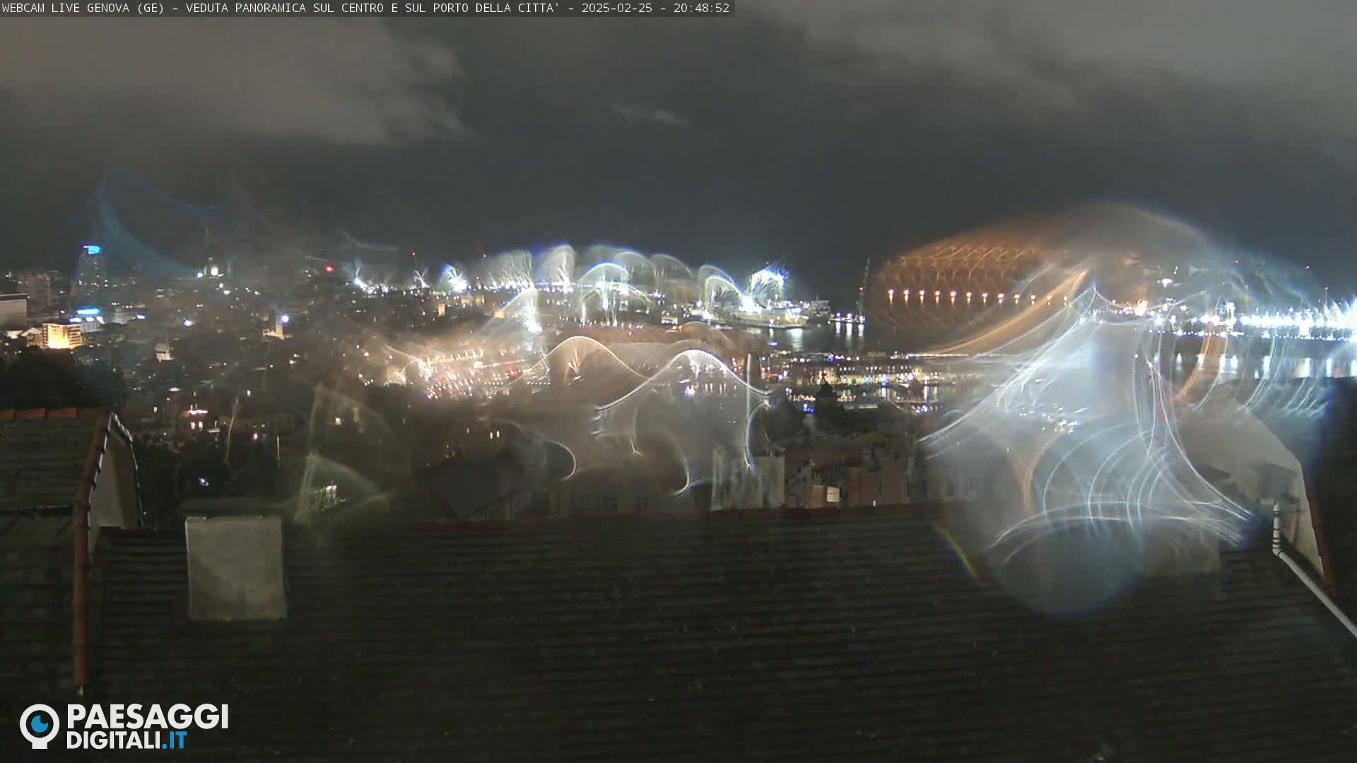 A nighttime view of a city harbor with a light show, viewed from a rooftop under a cloudy sky.