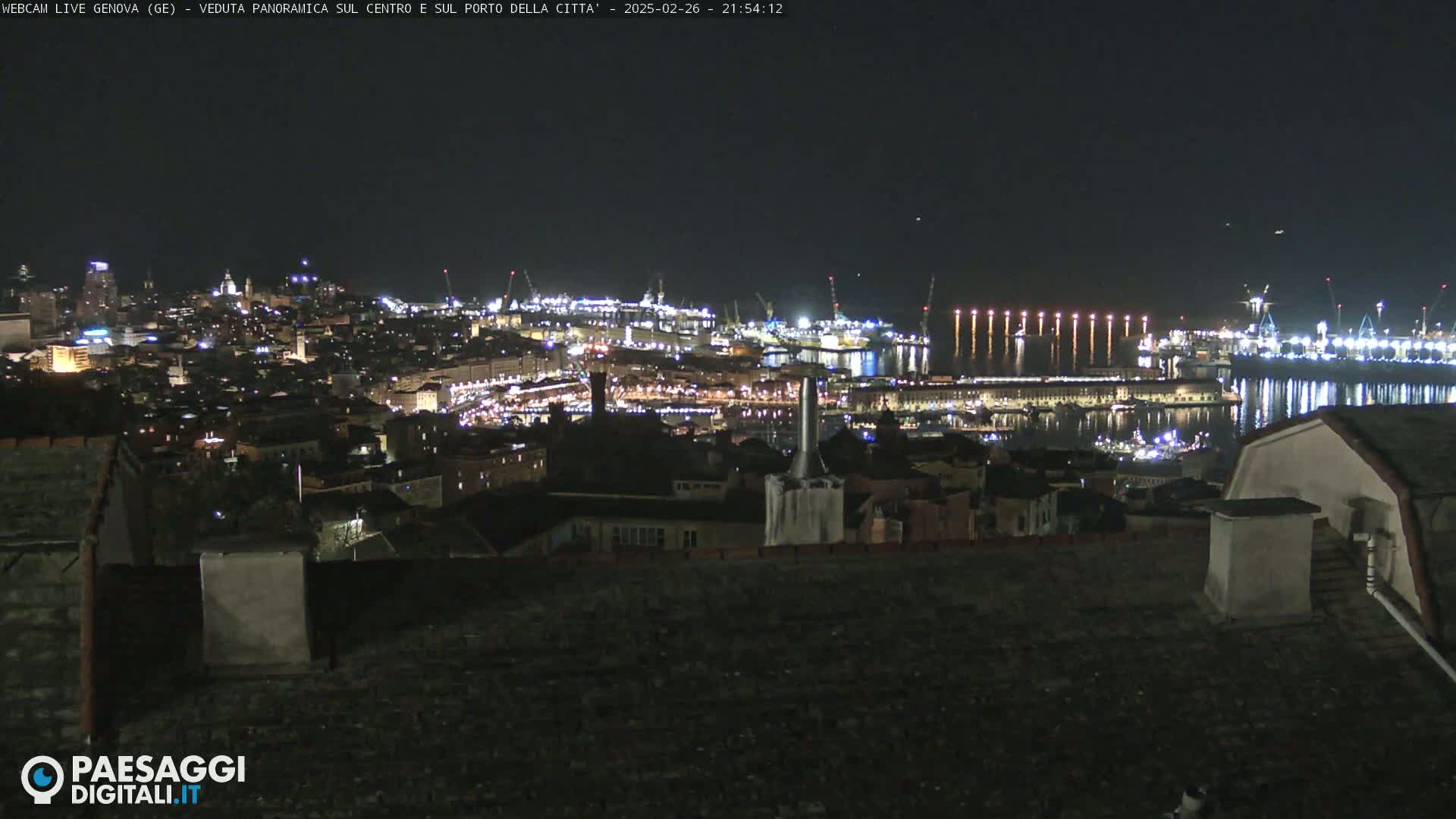 A nighttime panoramic view of a brightly lit city and harbor, with calm water reflecting the lights.