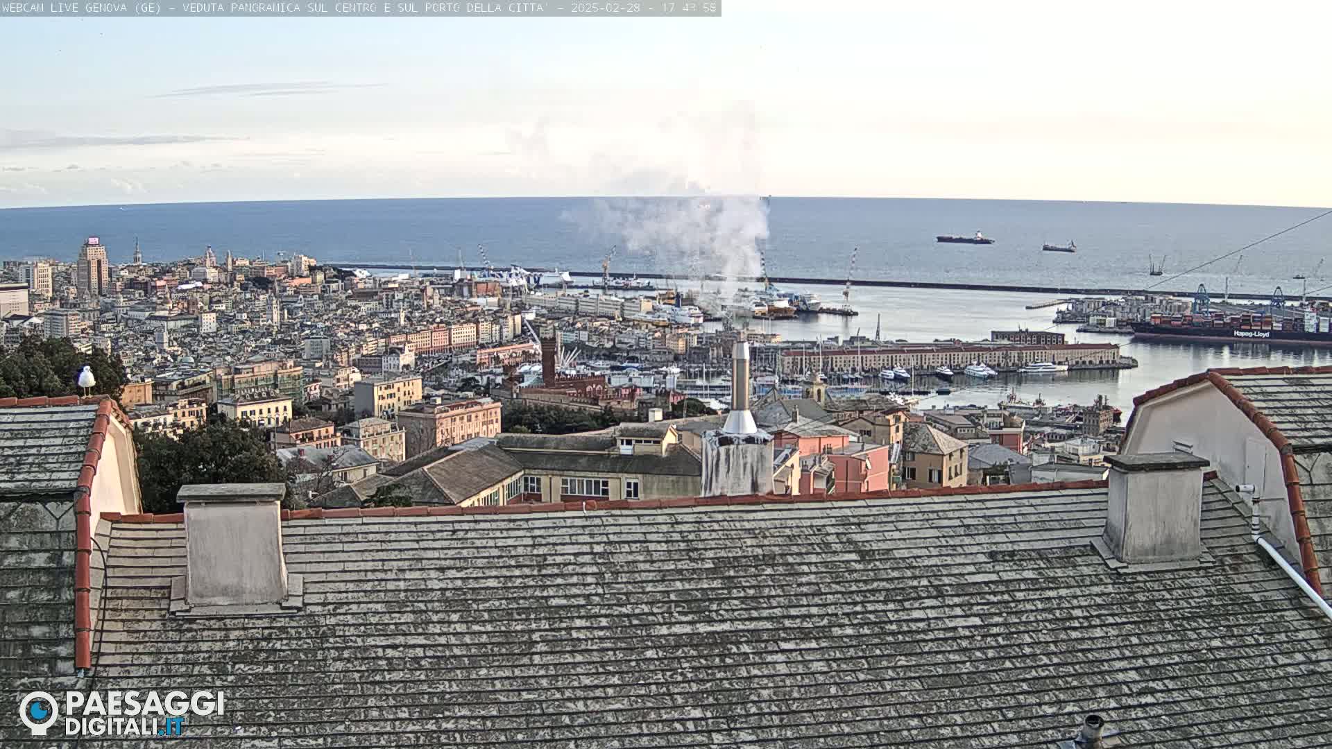 A panoramic view of a city with a harbor and ocean in the background on a mostly clear day; smoke is visible from a chimney in the foreground.