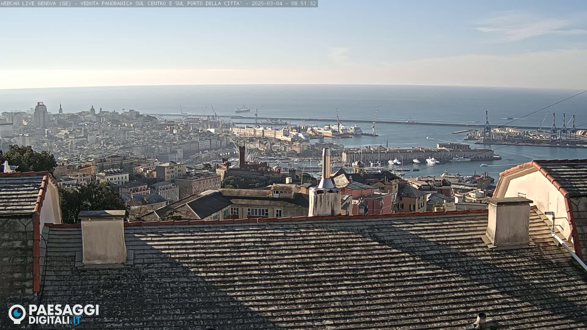 A panoramic view of a city and harbor on a sunny day, seen from a rooftop with a gray tiled roof.